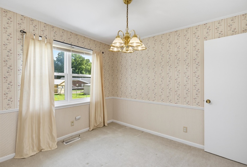 Carpeted interior space featuring a double-hung window, a brass-tone chandelier, and light-toned wallpaper with a floral pattern and a geometric wainscoting