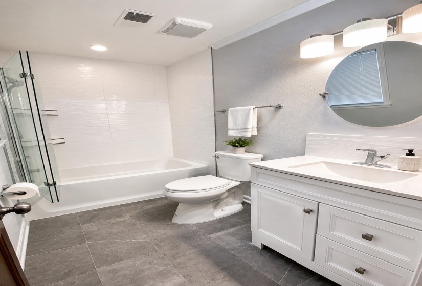 Full bathroom featuring a white vanity with integrated sink, a rectangular white tub with glass shower enclosure, and grey rectangular floor tiling