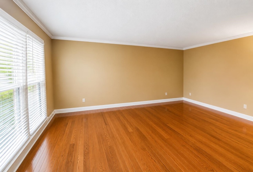 Spacious room featuring rich wood-finish flooring, neutral wall tones, white trim, and a large window with horizontal blinds