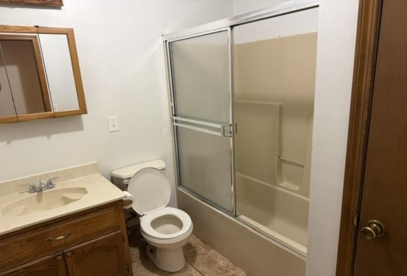 Full bathroom featuring a wood-finish vanity with an integrated sink, a commode, and a tub/shower combination with sliding frosted glass doors