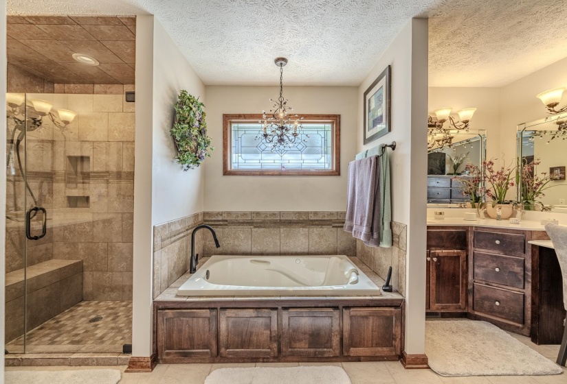 Spacious bathroom featuring a built-in jetted tub with wood-finish paneling, a large walk-in shower with tiled walls and a bench, and a dual vanity with dark wood cabinetry and individual mirrors