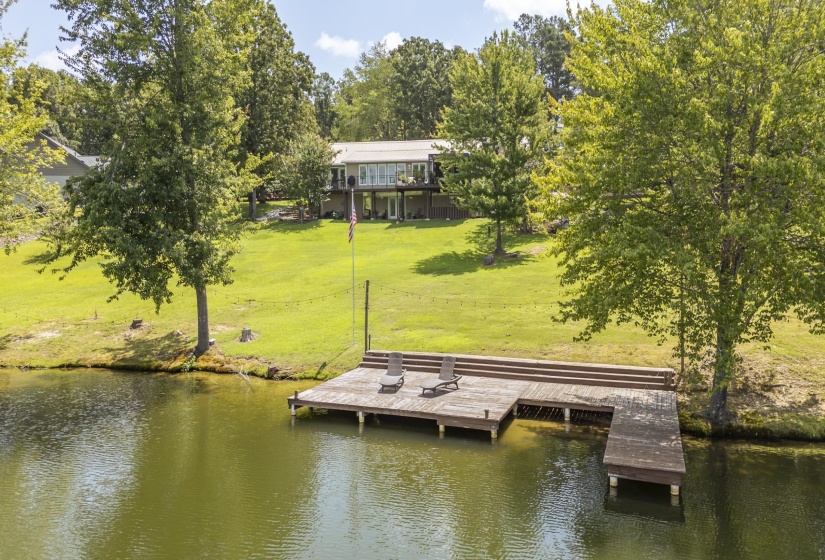 Expansive waterfront property featuring a large wooden dock, lush green lawn, mature trees, and a two-story residence with an upper-level balcony