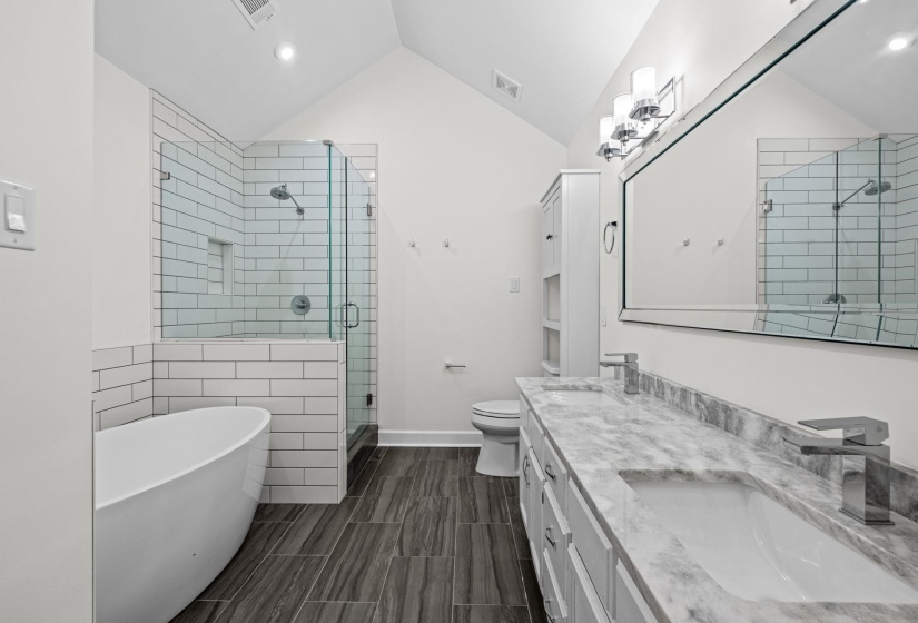 Modern bathroom featuring a freestanding soaking tub, a walk-in shower with subway tile surround and glass enclosure, a dual vanity with stone countertop and integrated sinks, wood-finish flooring, and a vaulted ceiling