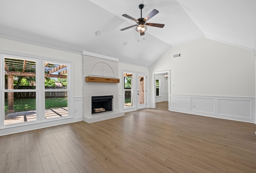 Spacious living area featuring vaulted ceilings, wood-finish flooring, a white brick fireplace with a wood mantle, and multiple windows providing outdoor access and views of a wood pergola