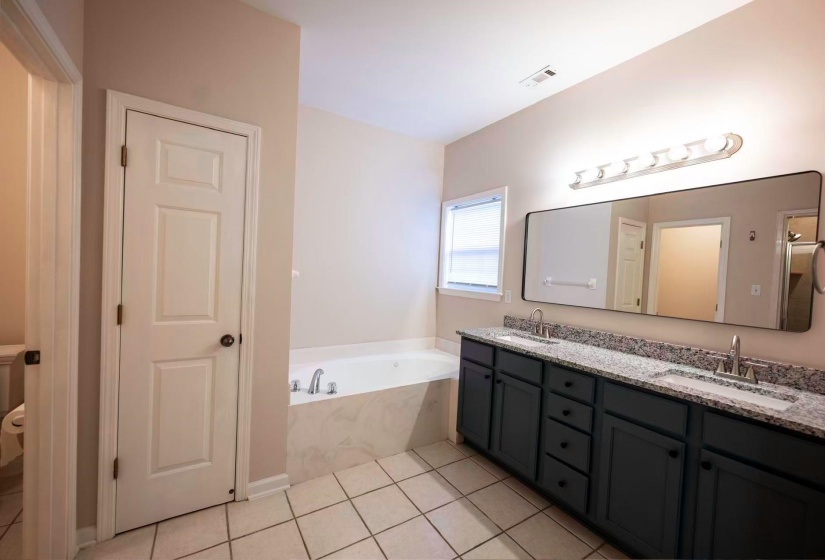 Bathroom featuring a dual vanity with a granite countertop, undermount sinks, a large wall-mounted mirror, a jetted tub, and tile flooring