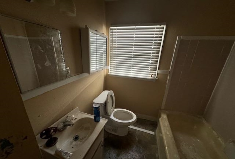Bathroom featuring a single window with blinds, a wall-mounted mirror, a built-in medicine cabinet, a toilet, a vanity with an integrated sink, and a bathtub with tile surround