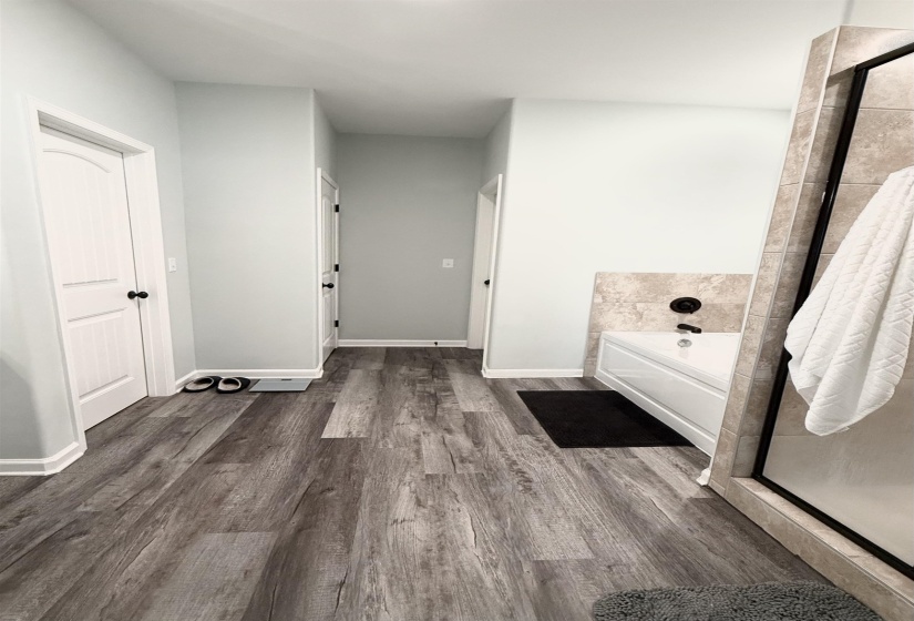 Spacious bathroom featuring wood-finish flooring, light grey walls, a built-in bathtub with a tiled surround, a glass-enclosed shower, and white interior doors with dark hardware