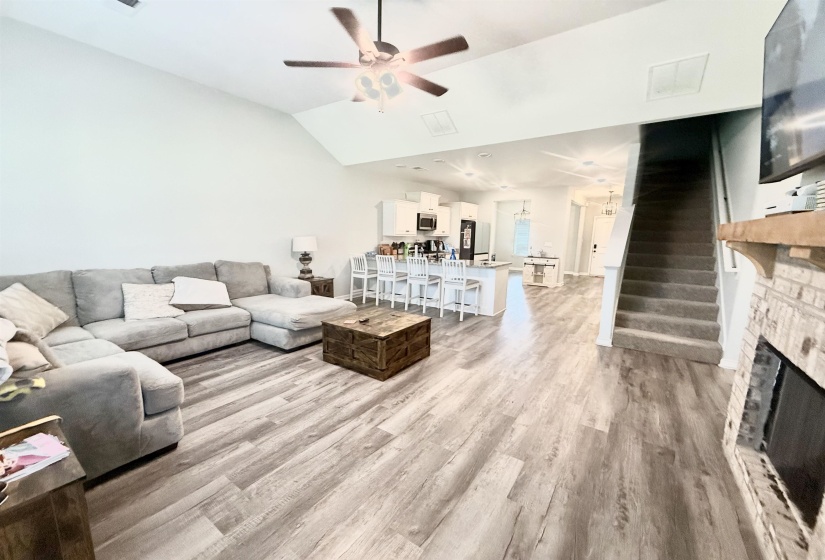 Open-concept living area with vaulted ceilings, wood-finish flooring, a ceiling fan, and a fireplace with brick surround and wood mantel