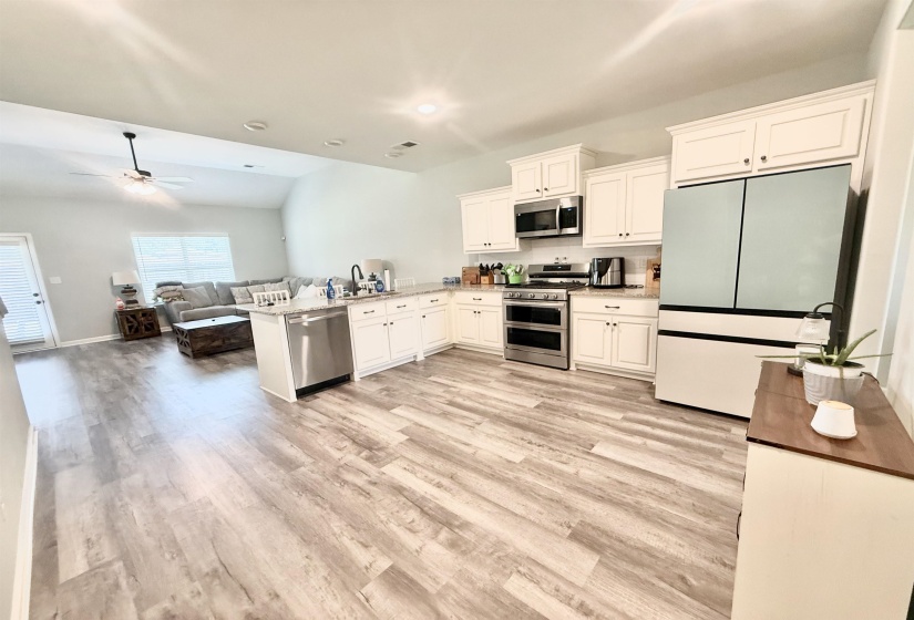 Open-concept kitchen featuring wood-finish flooring, white cabinetry with crown molding, stainless steel appliances, a white subway tile backsplash, and a breakfast bar with a stainless steel sink