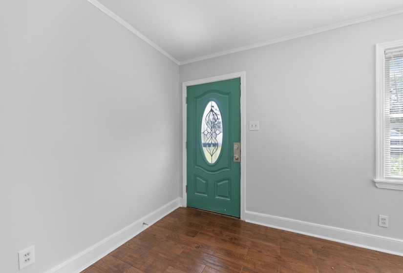 Entryway featuring a green front door with an oval leaded glass insert, wood-finish flooring, light gray walls, white trim, and a window with horizontal blinds