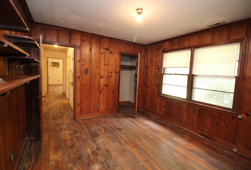 Room featuring wood-paneled walls, wood-finish flooring, built-in shelving, two windows with roller shades, and a ceiling-mounted light fixture