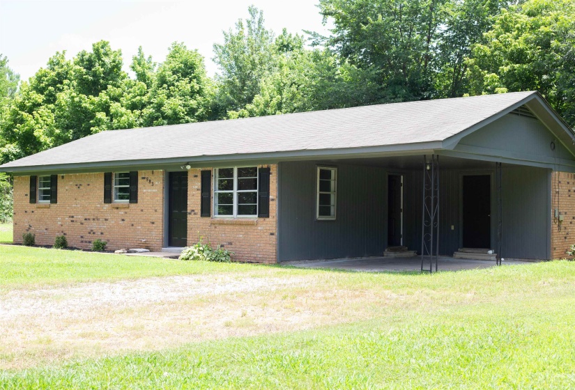 Brick and siding exterior featuring a carport with decorative metal posts, multiple windows with dark shutters, and a gravel driveway