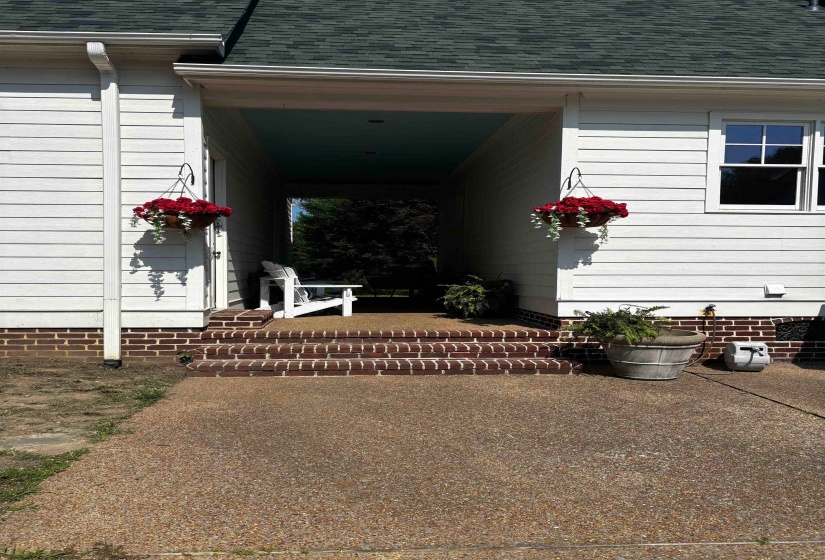 Covered walkway featuring white siding, a dark green shingle roof, brick steps, and exposed aggregate concrete paving