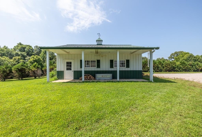 Two-tone white and green outbuilding featuring a covered porch, metal roof, cupola with weather vane, and a concrete patio