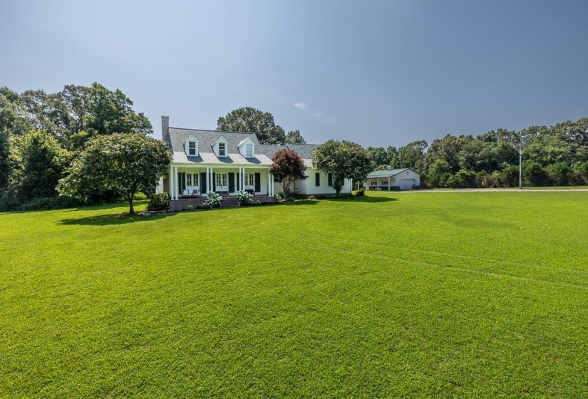 Expansive green lawn with a white colonial-style home featuring a covered front porch with white columns, dark shutters, and three dormer windows