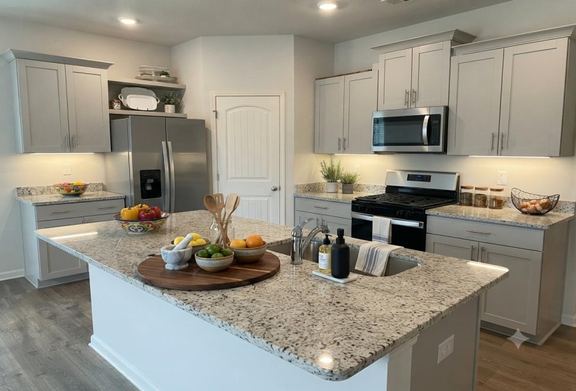 Kitchen featuring an island with an undermount sink, granite countertops, gray shaker-style cabinetry, stainless steel appliances, and wood-finish flooring
