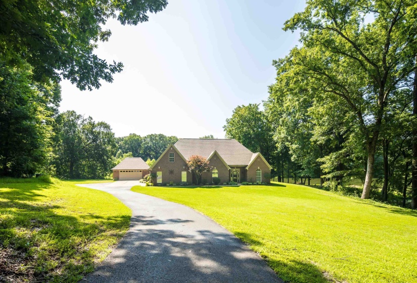 Brick exterior residence with an attached garage, featuring a long paved driveway, surrounded by mature trees and manicured lawns