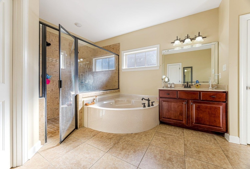 Bathroom featuring a corner soaking tub, a glass-enclosed shower with tiled walls, a single vanity with a stone countertop, a large mirror, and tiled flooring