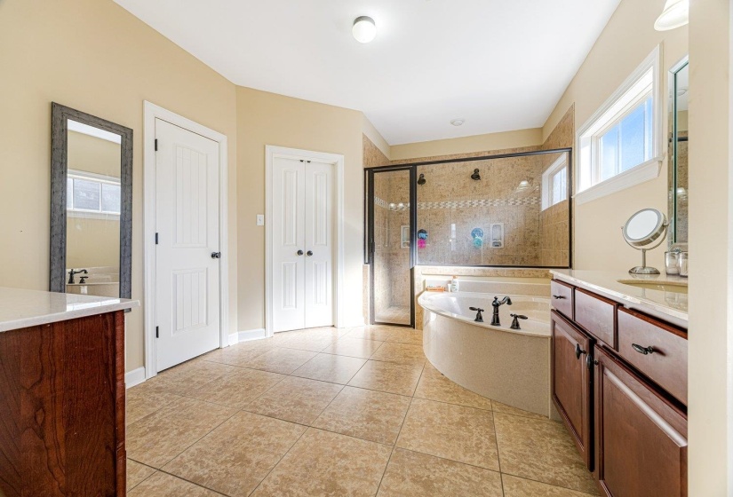 Bathroom featuring a large corner soaking tub, a tiled walk-in shower with a glass enclosure, dual sink vanity with dark wood cabinetry, and neutral-toned floor tiling
