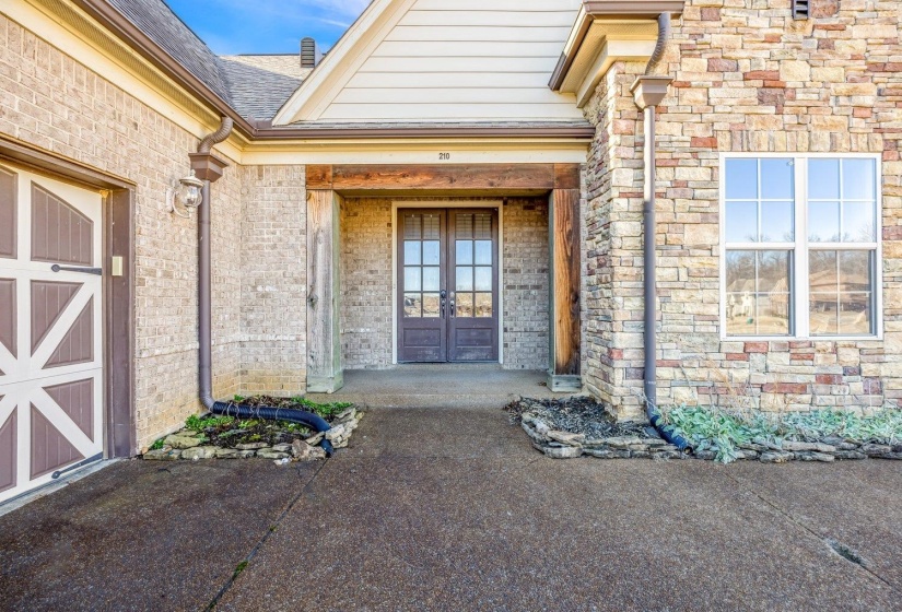 Front facade featuring a stone and brick exterior, double-door entry with glass panels, a covered porch with wood-finish columns, a decorative garage door, and a multi-pane window