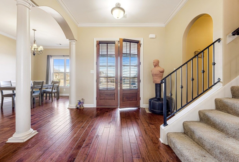 Entry foyer featuring rich wood-finish flooring, a carpeted staircase with wrought iron railing, decorative columns, and an arched doorway