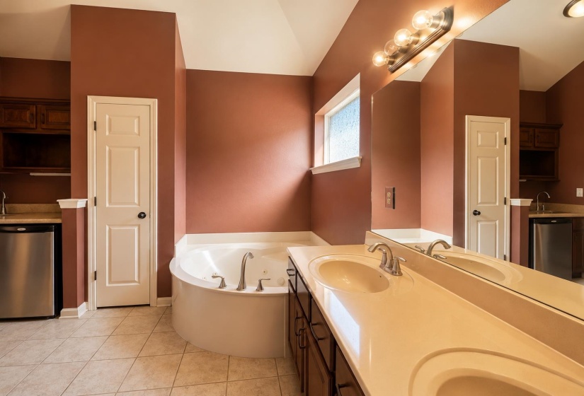 Bathroom featuring a built-in corner soaking tub, double vanity with integrated sinks, and a separate wet bar area with cabinetry and a compact refrigerator