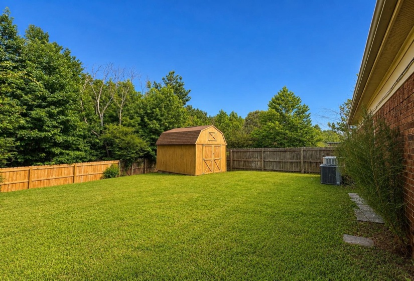 Expansive green lawn featuring a wooden storage shed, full privacy fencing, mature perimeter trees, and a brick exterior