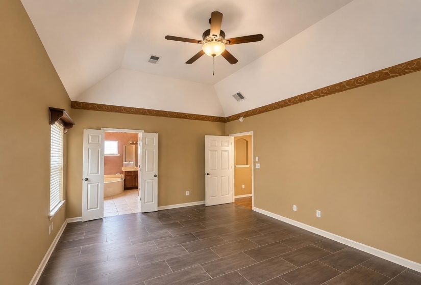 Spacious primary bedroom featuring vaulted ceilings, a ceiling fan with integrated lighting, wood-finish tile flooring, and neutral wall tones with a decorative border