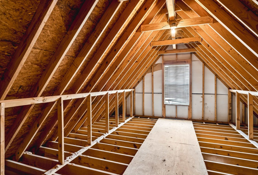 Unfinished attic space featuring exposed wood framing, a pitched roofline, partial wall sheathing, and a window with blinds