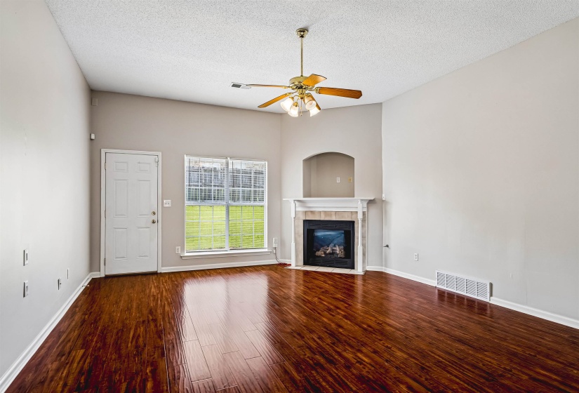 Living area featuring wood-finish flooring, a fireplace with a mantel and arched niche, an exterior door, and a window overlooking green space