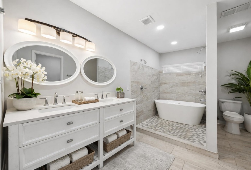 Contemporary bathroom featuring a dual vanity with white cabinetry and countertop, oval mirrors, a freestanding soaking tub, and a separate shower area with tiled walls and flooring