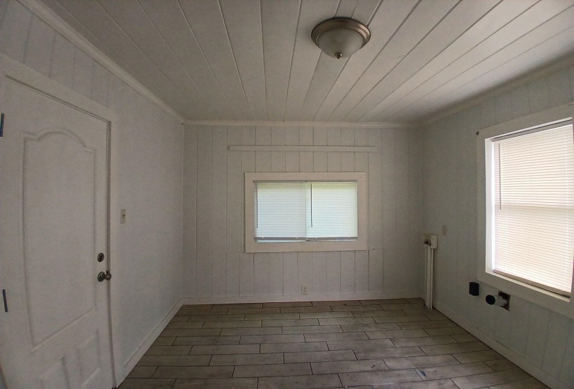 Interior room featuring plank-style walls, wood-finish flooring, two windows with blinds, a ceiling-mounted light fixture, and a paneled door