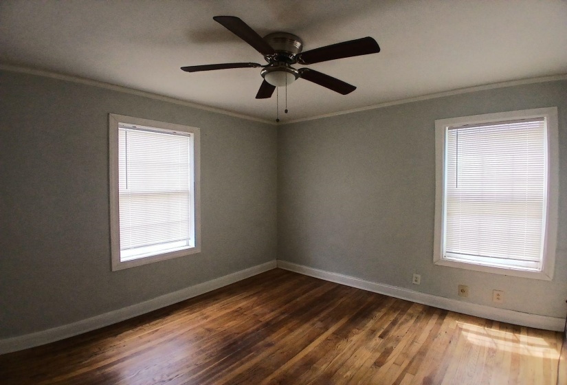 Wood-finish flooring throughout, featuring two windows with horizontal blinds, a ceiling fan with integrated lighting, crown molding, and baseboards