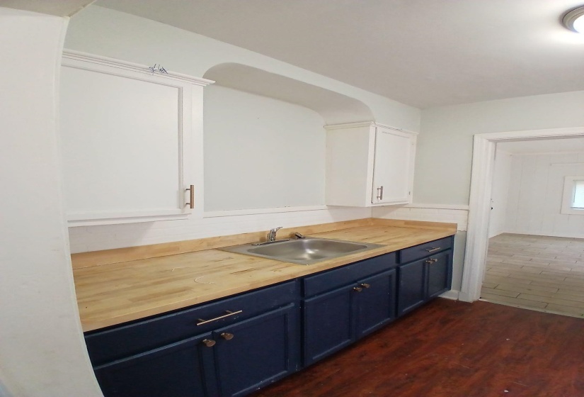 Kitchenette featuring a stainless steel sink, wood-finish countertop, navy lower cabinetry with brass hardware, white upper cabinetry, and wood-finish flooring