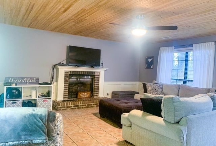 Living area featuring a brick fireplace with a white mantel, wood-finish ceiling, ceiling fan, tile flooring, and a large window with blinds and sheer curtains