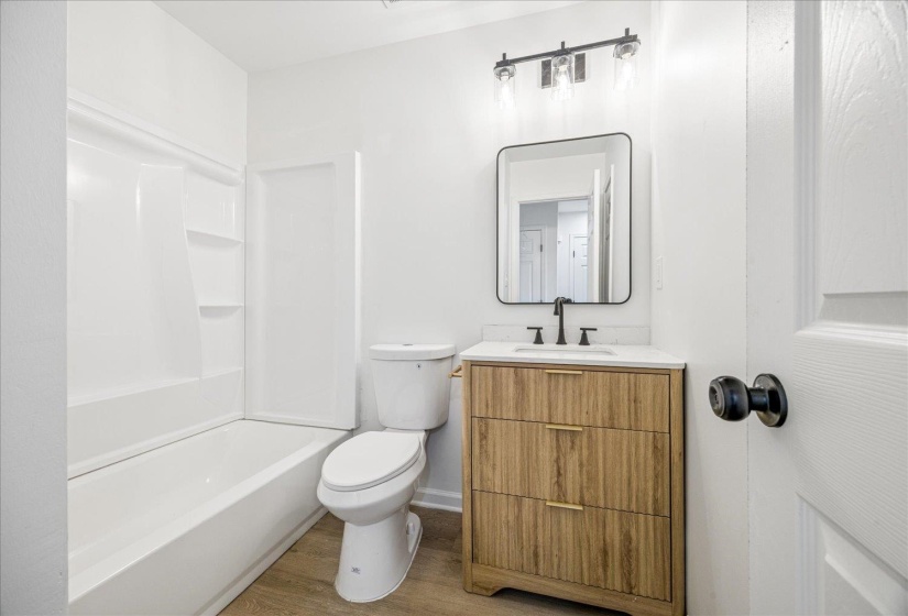 White bathroom featuring a single vanity with a wood-finish cabinet and gold-tone hardware, a white countertop, and a rectangular mirror with a black frame
