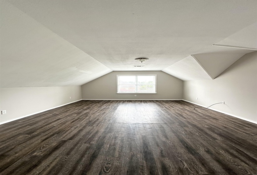 Spacious room featuring vaulted ceilings, a central window with natural light, wood-finish flooring, and neutral wall tones