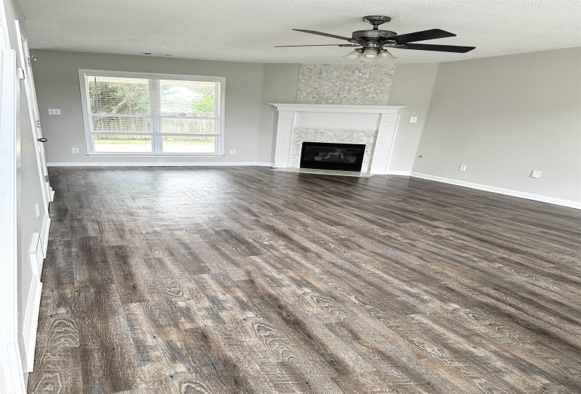 Spacious room featuring wood-finish flooring, a fireplace with a white mantel and tiled surround, a decorative tiled accent wall, and a ceiling fan with integrated lighting