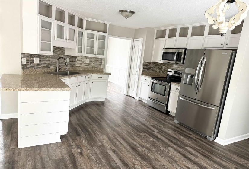 Kitchen featuring wood-finish flooring, white cabinetry with glass-front uppers, a mosaic tile backsplash, stainless steel appliances, and L-shaped countertops