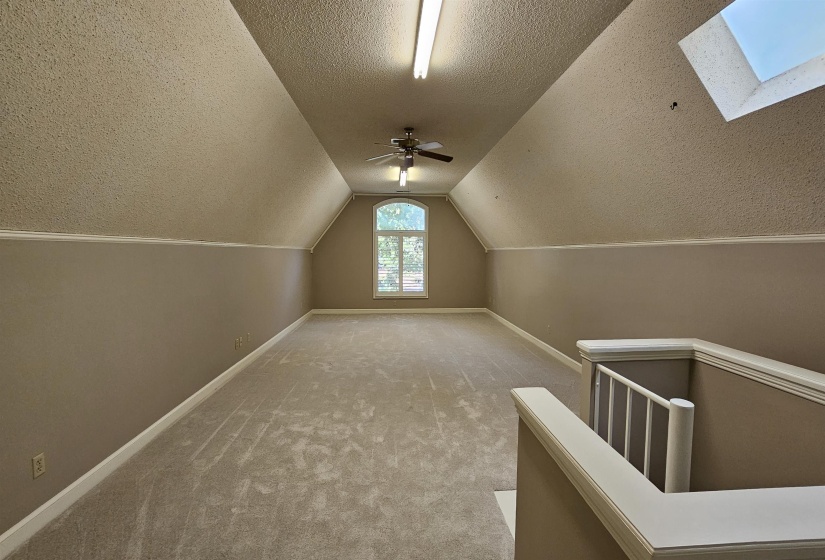 Spacious loft room featuring vaulted ceilings, neutral carpeting, a skylight, an arched window, and a ceiling fan