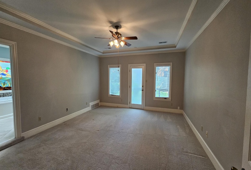 Spacious room featuring tray ceilings with crown molding, plush carpeting, ceiling fan with integrated lighting, and multiple windows and a glass-paneled door for abundant natural light