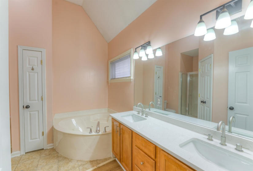 Bathroom featuring a corner soaking tub with deck-mounted fixtures, a dual vanity with light-toned countertops and undermount sinks, and a full-width mirror with a multi-bulb vanity light