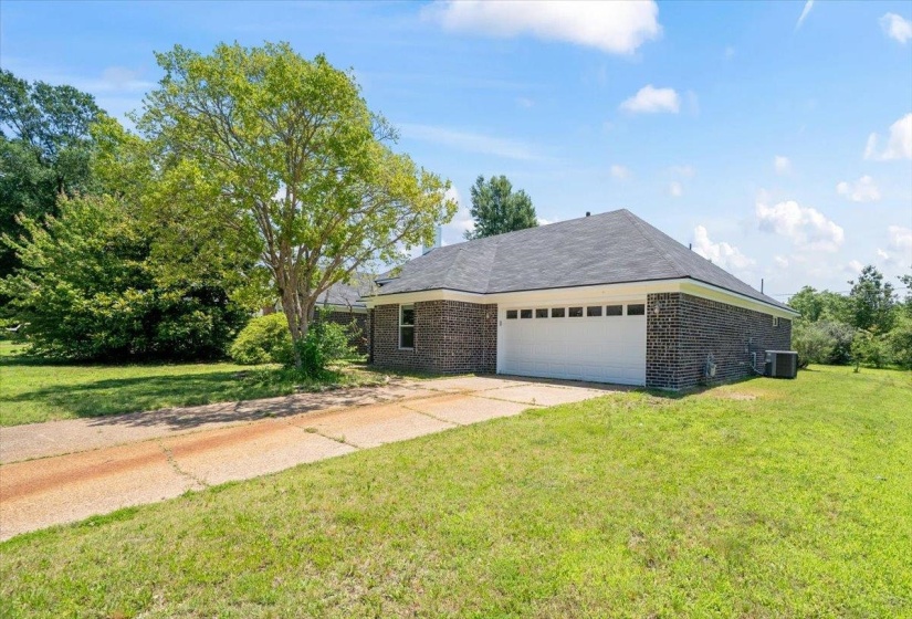 Dark brick exterior home featuring a two-car garage with a white door, a gray shingled roof, a concrete driveway, and a large mature tree