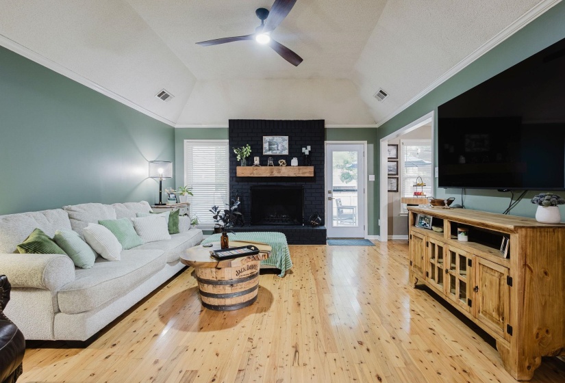 Vaulted ceiling living area featuring wood-finish flooring, a black brick fireplace with a wood mantel, and crown molding