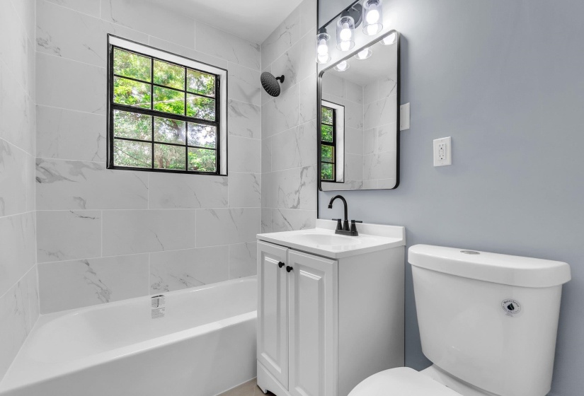 Bathroom featuring white subway tile with subtle veining, a large soaking tub, a single-pane window with black framing, a vanity with a white countertop and black hardware, and a wall-mounted mirror with integrated lighting