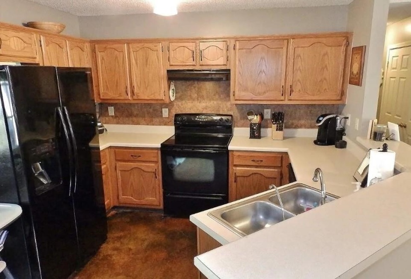 Kitchen with wood-finish cabinetry, solid surface countertops, a double basin stainless steel sink, and a full suite of black appliances, including a side-by-side refrigerator with dispenser