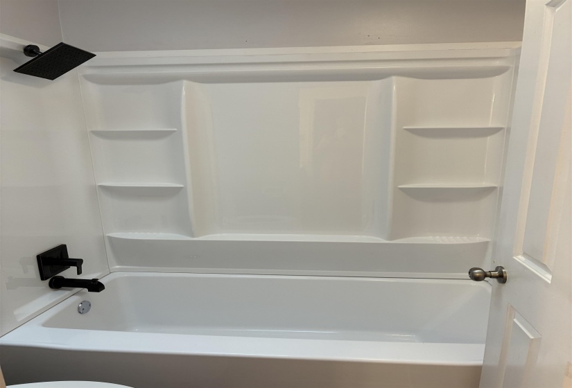 White bathtub and shower surround featuring integrated shelving, a matte black rainfall showerhead, and coordinating matte black tub filler and controls