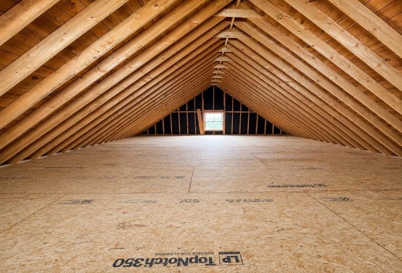 Expansive attic space featuring exposed wood rafter beams, durable subflooring, and a framed window opening