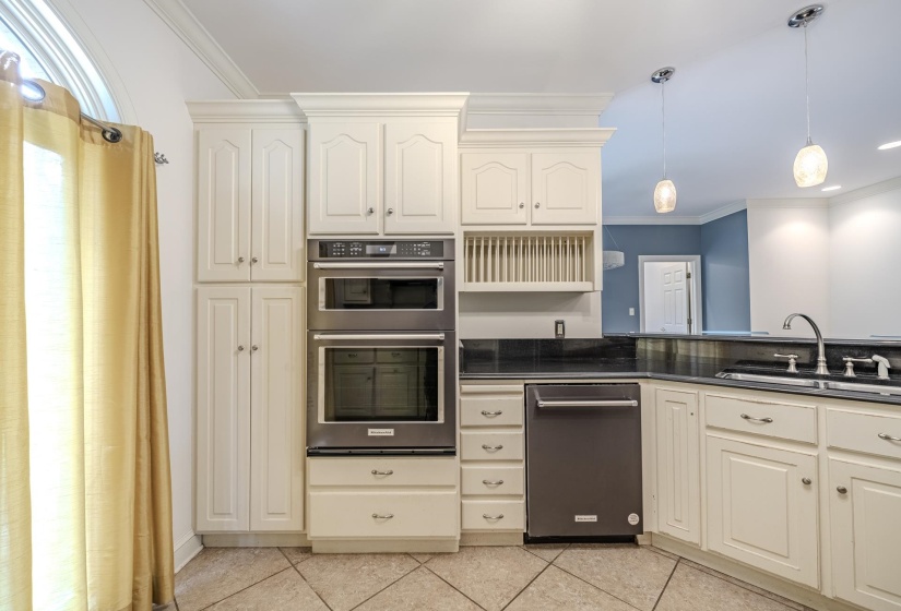 Kitchen featuring cream-painted cabinetry with crown molding, double wall ovens, a stainless steel dishwasher, a dark countertop, and a double basin sink with a chrome faucet