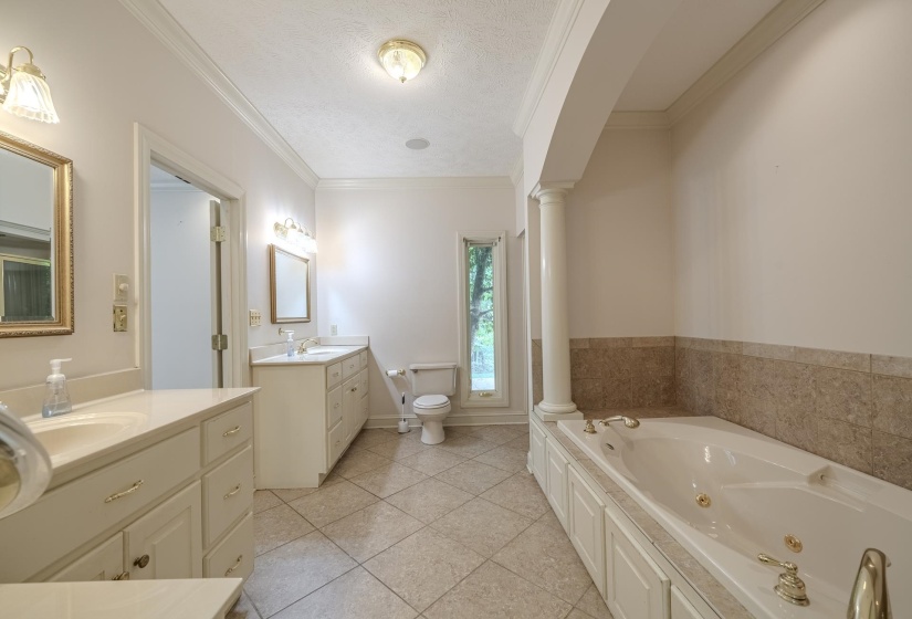 Spacious bathroom featuring two separate vanity areas, a built-in jetted tub with gold-tone fixtures, a tiled half-wall, and a tall window